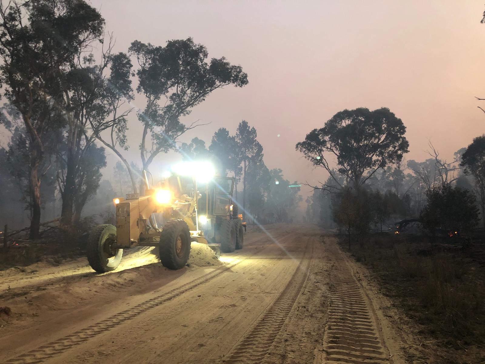 Grader cutting a containment line at dusk during bushfires with smoke in the air