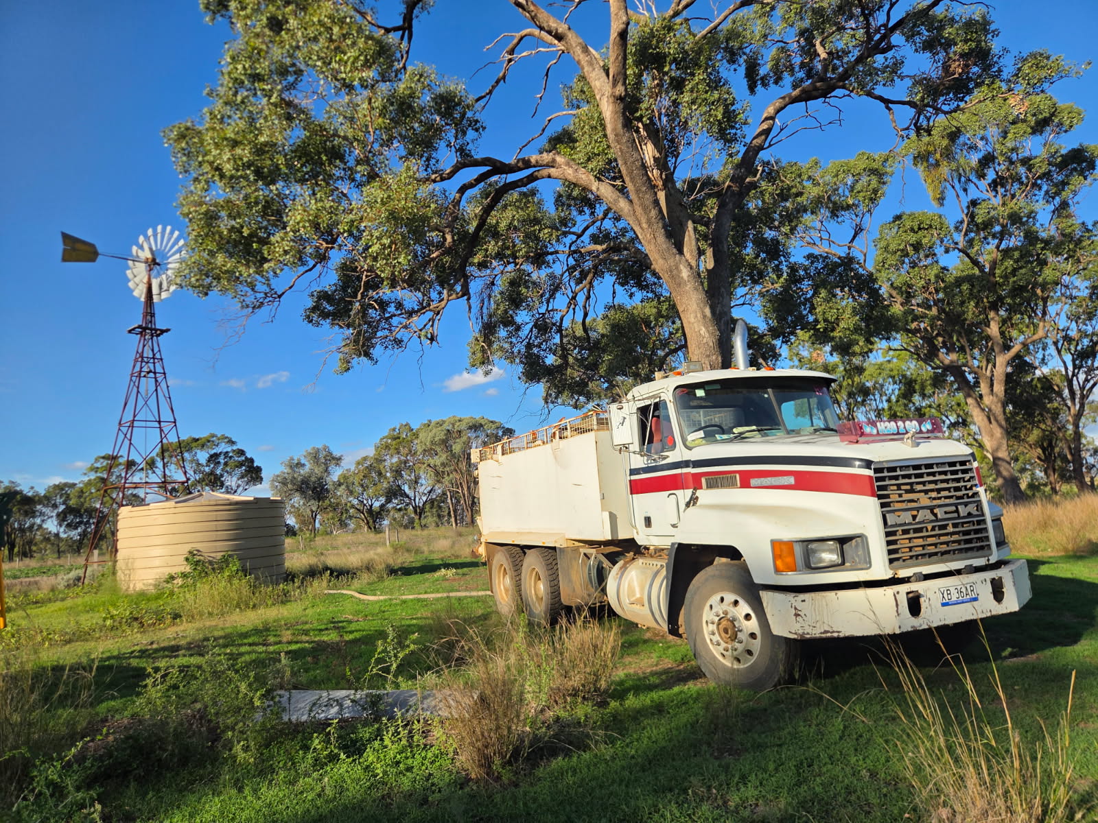 Mack water cart truck on a rural property at golden hour with windmill and water tank in the background